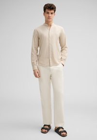 Beige overhemd met lange mouwen en knoopsluiting, voorzien van een Mandarin kraag, gecombineerd met off-white broeken en zwarte sandalen, met een relaxte pasvorm en een gestructureerde stof.