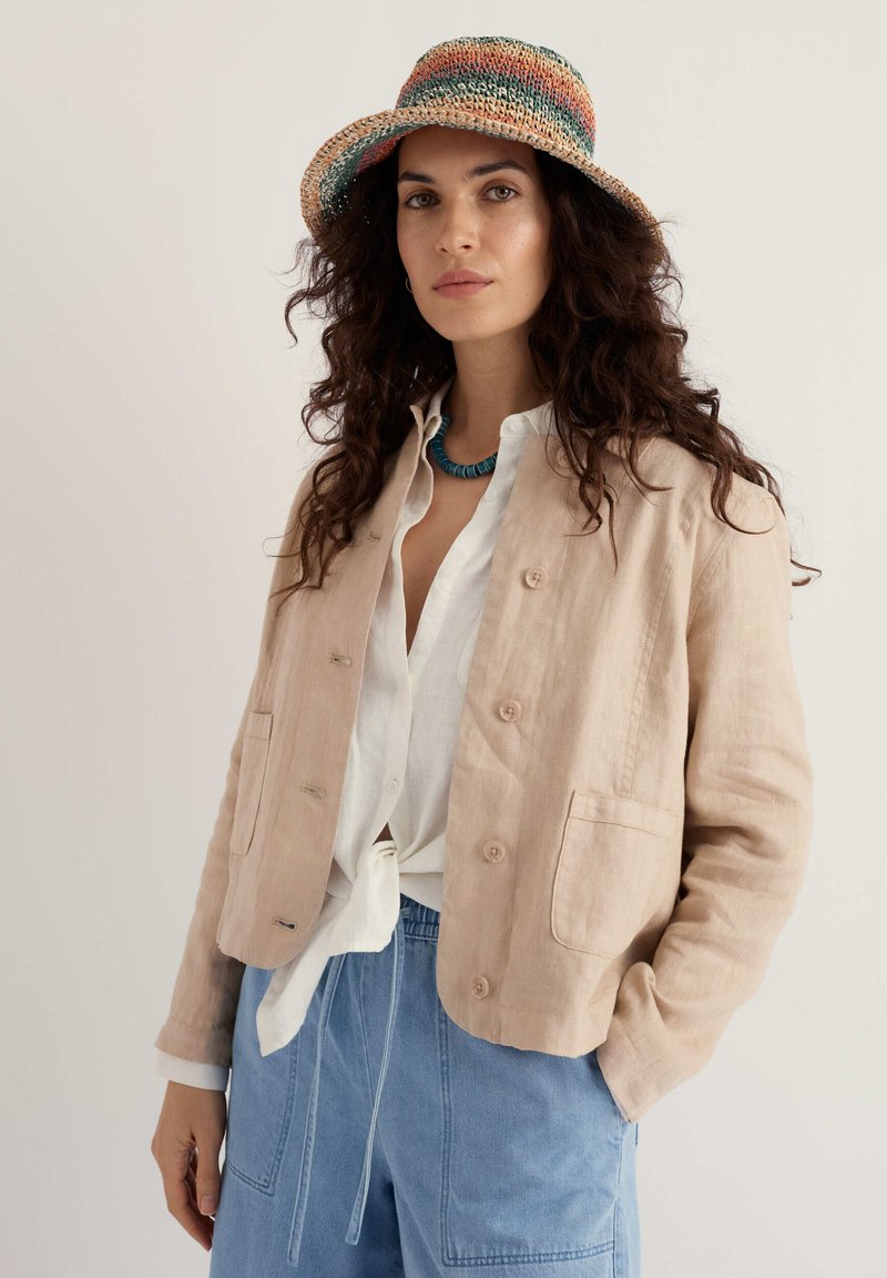 Femme aux longs cheveux bouclés portant un bonnet tricoté multicolore, une veste beige boutonnée, une chemise blanche nouée, un pantalon bleu et un collier turquoise.
