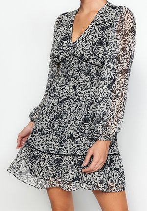 Frau trägt ein schwarz-weiß gemustertes, langärmeliges Kleid mit V-Ausschnitt und einem Rüschenabschluss, steht vor einem schlichten weißen Hintergrund.