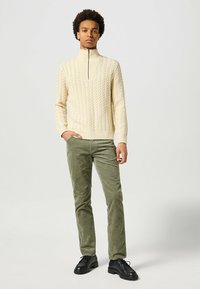Jasno beżowy sweter w warkocze z wysokim kołnierzem, zapinany na zamek, w połączeniu z oliwkowymi spodniami z materiału sztruksowego i czarnymi butami na sznurowadła.