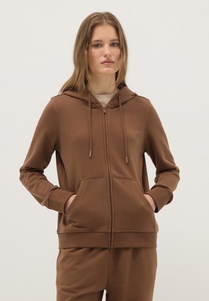 Mujer joven vistiendo una sudadera marrón con cremallera y cordones, y pantalones a juego, de pie con las manos en los bolsillos de la sudadera, fondo neutro.