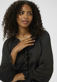 Blusa nera con colletto arricciato, maniche arricciate e texture liscia. La vestibilità è comoda, con dettagli sottili ai polsini.