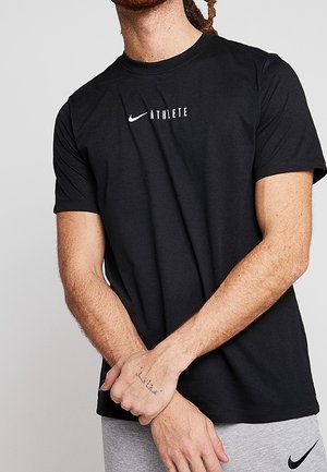 Mand iført sort Nike t-shirt med "ATHLETE" tekst og grå Nike bukser, stående med armene krydset foran.