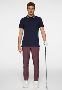 Marinblå polo-shirt med randig krage, i kombination med burgundy byxor. Modell som håller en golfklubba, iförd vita skor och en golfhandske.