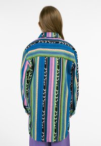 Pestrá pruhovaná košeľa s modrými, ružovými, žltými a zelenými vzormi a čiernymi akcentmi. Voľný strih a dlhé rukávy. Textúrovaná tkanina.