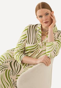 Langärmliges Kleid in Creme mit grünen wellenförmigen Streifen, ausgestattet mit einem Kragen und einem Knopfdetail, kombiniert mit einer auffälligen Halskette. Glatte Textur.