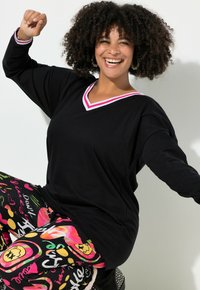 Musta pitkähihaispaita, jossa on punavalkoinen V-pääntie, yhdistetty värikkäisiin kuvioituihin pyjamahousuihin, joissa on sydämiä ja graafisia kuvioita.