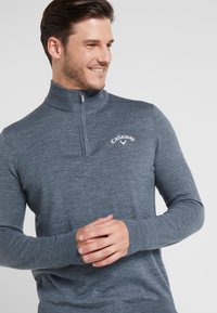 Pullover cinza com fecho na gola, feito de um material de malha texturizado, apresentando um logótipo no peito e mangas longas com punhos canelados.