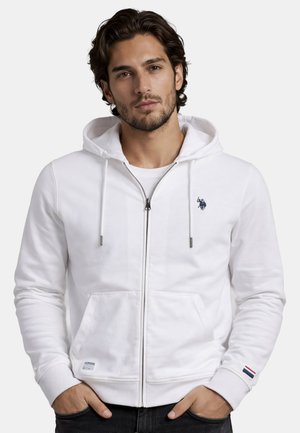 Man wearing white zip-up hoodie with navy logo on chest and small flag detail on sleeve, hands in pockets, neutral background.