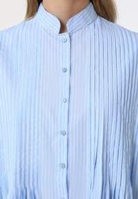 Chemise bleu clair avec col mao, rayures verticales et patte de boutonnage. Le tissu est lisse avec de petites plis subtils partout.
