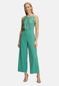 Grön jumpsuit med volangaxlar, vida ben och åtsittande midja. Kombinerad med strappy metalliska klackar. Slät tygstruktur.