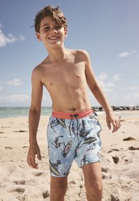 Smiling boy wearing blue swim shorts with skeleton surfer prints, standing on a sandy beach under a clear sky.