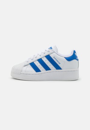 Baskets blanches Adidas Superstar avec des rayures et accents bleus, lacées, montrées en profil sur un fond gris uni.