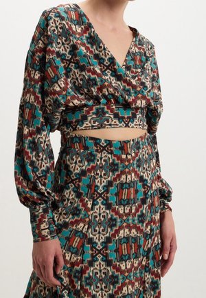 Femme portant un crop top à manches longues assorti et une jupe taille haute avec un motif géométrique coloré en bleu sarcelle, marron, crème et noir.