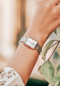 Reloj rectangular plateado con cara blanca, números romanos y una correa de metal texturizada. Mano descansando cerca de hojas verdes.