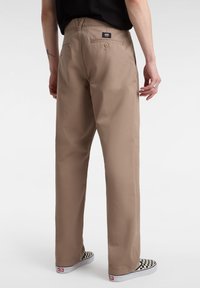 Pantaloni a gamba larga tan con una vestibilità casual, dotati di tasche posteriori, chiusura a bottone e etichetta, abbinati a scarpe slip-on a scacchi.