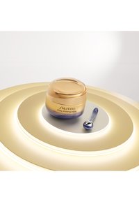 Shiseido Vital Perfection ögonkräm i en rund glasburk med en guld- och ombre-finish. Åtföljd av en blå appliceringssked.