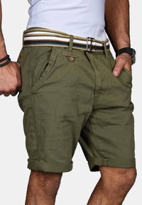 Mann trägt olivgrüne hochgekrempelte Shorts mit gestreiftem Stoffgürtel, weißem Hemd, das eingesteckt ist, schwarze Armbanduhr und schwarzes Perlenarmband.
