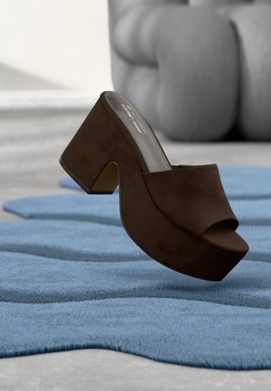 Mule à plateforme en daim marron exposée sur un tapis bleu texturé avec une sculpture abstraite grise en arrière-plan.