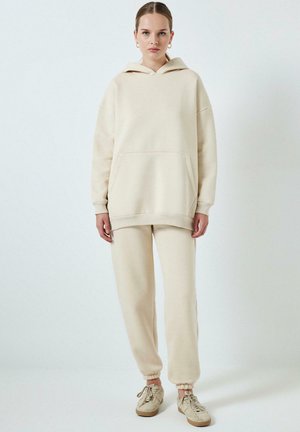 Sudadera beige y pantalones de chándal a juego, confeccionados en tejido suave; la sudadera cuenta con un bolsillo frontal y puños anchos, mientras que los pantalones tienen tobillos elásticos.