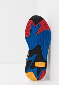 Semelle de chaussure avec un motif multicolore incluant du rouge, bleu, jaune et noir en caoutchouc, texturée pour l'adhérence et la stabilité. Une étiquette de marque est visible.