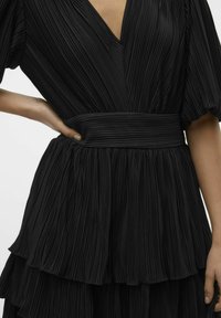 Czarna plisowana sukienka z głębokim dekoltem w kształcie litery V, szerokim paskiem w talii i warstwami falban. Wykonana z teksturowanej, lekko opadającej tkaniny.