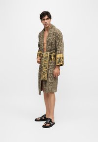 Άνδρας στέκεται φορώντας καφέ ρόμπα Versace με χρυσαφένιες μπαρόκ λεπτομέρειες και μαύρα σανδάλια σε λευκό φόντο.
