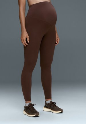 Personne enceinte portant des leggings de maternité marron et des baskets foncées, debout devant un fond gris.