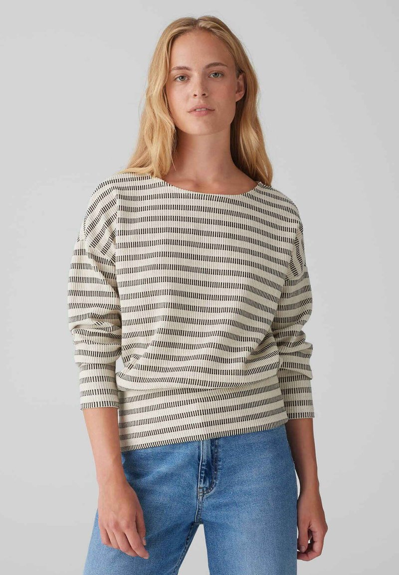 Striped long-sleeve top in cream and black, made from soft fabric. Features a round neckline and relaxed fit. Paired with blue jeans.