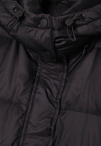 Veste matelassée noire avec col montant, cordon de serrage, boutons-pression et texture lisse. Caractérisée par des panneaux cousus et une poche zippée.