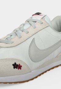 Λευκό sneaker Nike με γκρι swoosh, ανοιχτό γκρι κορδόνια, υφασμένο περίγραμμα και μικρά μαύρα και κόκκινα λουλουδένια σχέδια κοντά στη σόλα.
