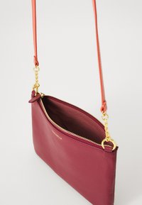 Mala crossbody iz burgundske kože z zlatim zadrgo in povezovalci za verigo, z oranžnim trakom in blagovno znamko "Coccinelle" spredaj.