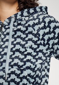 Sammet zip-up hoodie i ljusblå med mörkblå textmönster. Funktioner inkluderar broderad logotyp, dragsko och en mjuk textur.