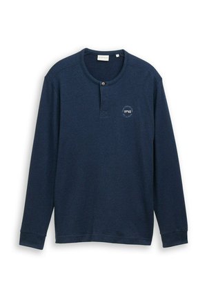 Camisa henley de manga larga azul marino con un botón cerca del cuello redondo y un pequeño texto impreso en círculo en el lado izquierdo del pecho.