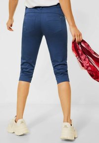 Námořnické modré capri kalhoty vyrobené z elastického materiálu s hladkou texturou, s zadními kapsami a přiléhavým střihem. Drží šátek v ruce.