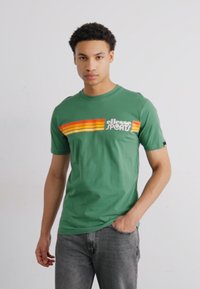 Grøn kortærmet t-shirt med lodrette striber i orange, gul og rød, med "ellesse SPORTS" logo, lavet af blødt bomuldsmateriale.