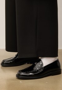 Fekete bőr loaferek fényes felülettel, fehér zoknival és fekete bő nadrággal párosítva, bézs padlón.