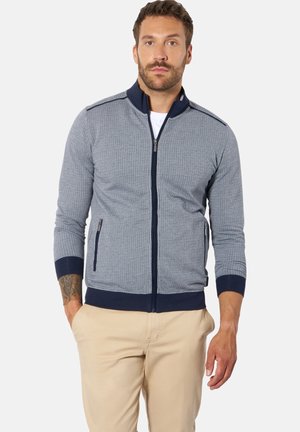 Mann steht mit den Händen in den Taschen, trägt eine graue und marineblaue, strukturierte Jacke mit Reißverschluss und beige Hose vor weißem Hintergrund.
