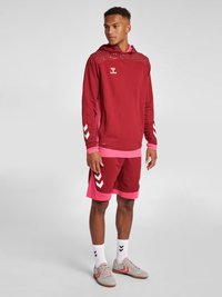 Röd atletisk hoodie med meshdetaljer och rosa accenter, tillsammans med röda shorts som har ett vitt chevronmönster. Grå sportskor och vita strumpor.