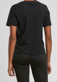 Achteraanzicht van een persoon in een eenvoudige zwarte T-shirt met korte mouwen en zwarte leggings tegen een effen grijze achtergrond.