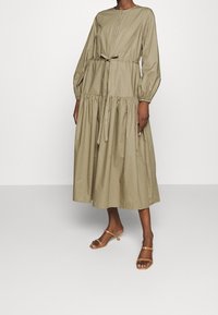 Donna che indossa un vestito midi beige con maniche lunghe, cintura arricciata in vita e sandali con tacco color nocciola, in piedi di fronte a uno sfondo semplice.