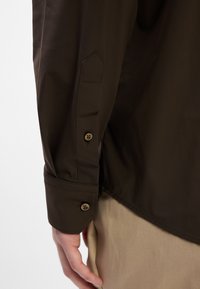 Camisa marrón de botonadura con un puño bien doblado y un diseño de mangas puntiagudas, con dos botones en el puño para el cierre.