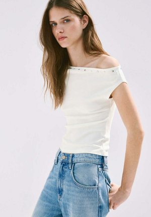 Jeune femme aux longs cheveux bruns portant un haut blanc épaules dénudées et un jean bleu taille haute, debout devant un fond clair uni.