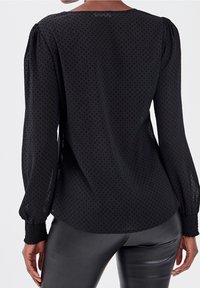 Musta läpikuultava pitkähihainen paita, jossa on pieniä pilkkuja, kootut hihansuut, mustien nahkahousujen kanssa, nähtynä takaa.