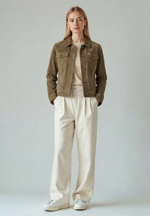 Ung kvinne står med hendene i lommene, iført olivengrønn semsket jakke, beige topp, vide hvite bukser og hvite sneakers mot ensfarget bakgrunn.