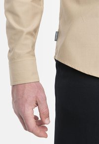 Camicia beige a maniche lunghe con polsini abbottonati, mostrata con una mano, abbinata a pantaloni scuri. Il tessuto appare liscio con una texture minima.