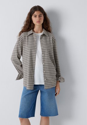 Woman with long hair wearing a loose checkered button-up shirt, white t-shirt, and blue knee-length denim shorts standing against a white wall.