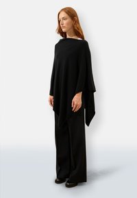 Czarny ponczo o luźnym, płynącym kroju, wykonane z miękkiego materiału. Charakteryzuje się jednolitą fakturą i asymetrycznym dołem, w zestawieniu z szerokimi czarnymi spodniami.