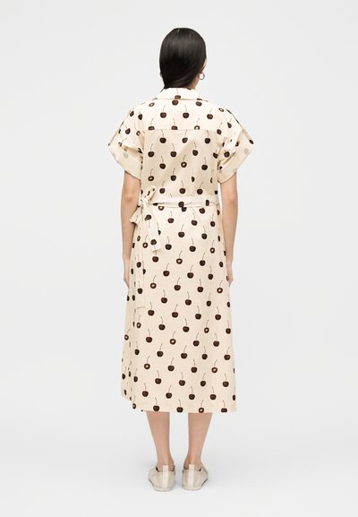 Γυναίκα με σκούρα μαλλιά που φοράει μπεζ midi φόρεμα με καφέ σχέδια κερασιών, κοντά μανίκια, ζώνη δεμένη και μπεζ παπούτσια, γυρισμένη με την πλάτη.
