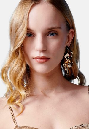 Una mujer con largo cabello rubio ondulado lleva un arete elegante y una blusa de estampado, mostrando una expresión suave contra un fondo claro.